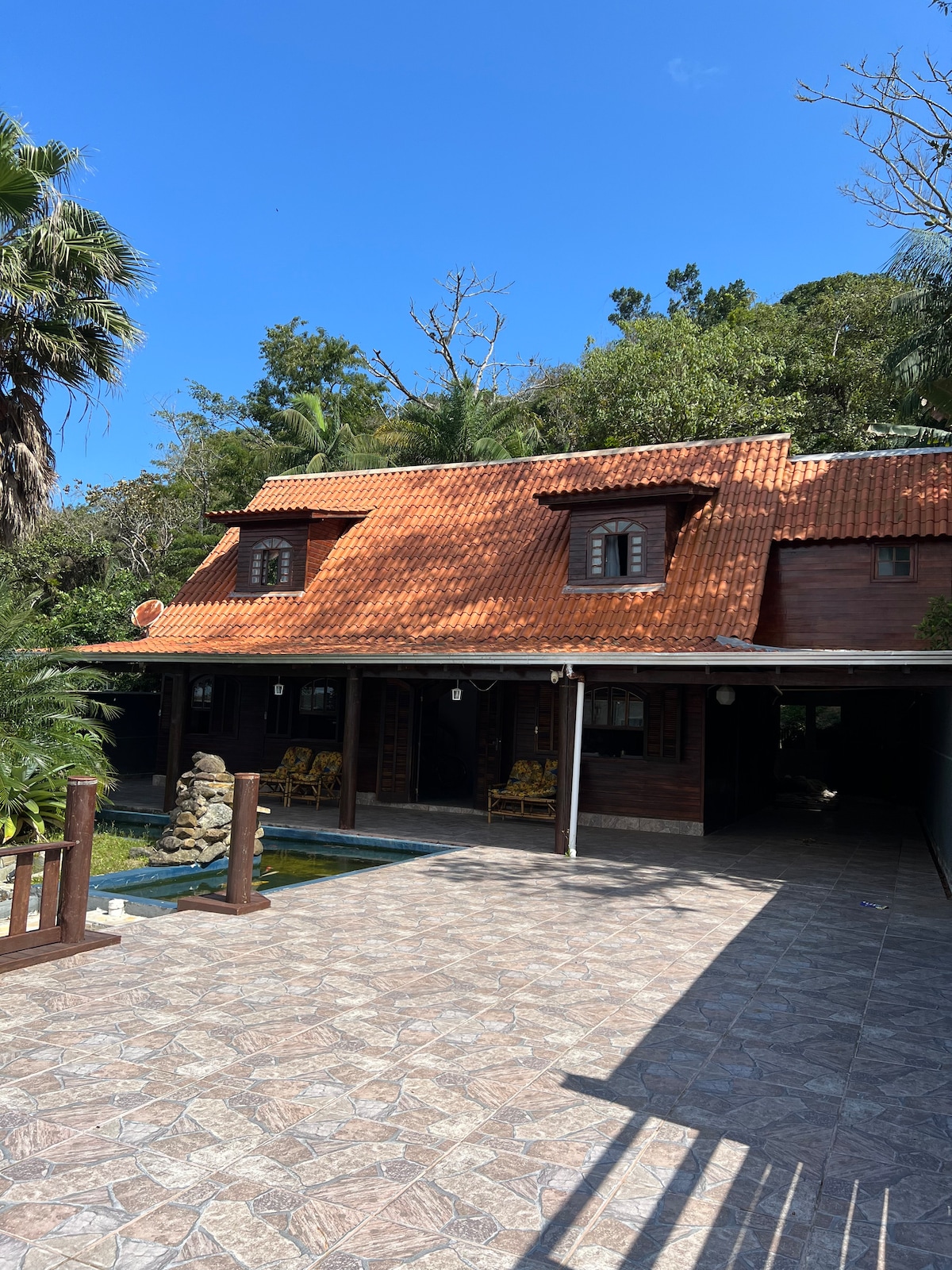 A charming wooden house is set against a backdrop of lush greenery. The red-tiled roof, flanked by large windows, complements the natural surroundings. A stone pathway leads to a pool area, inviting relaxation in the tranquil outdoor space.