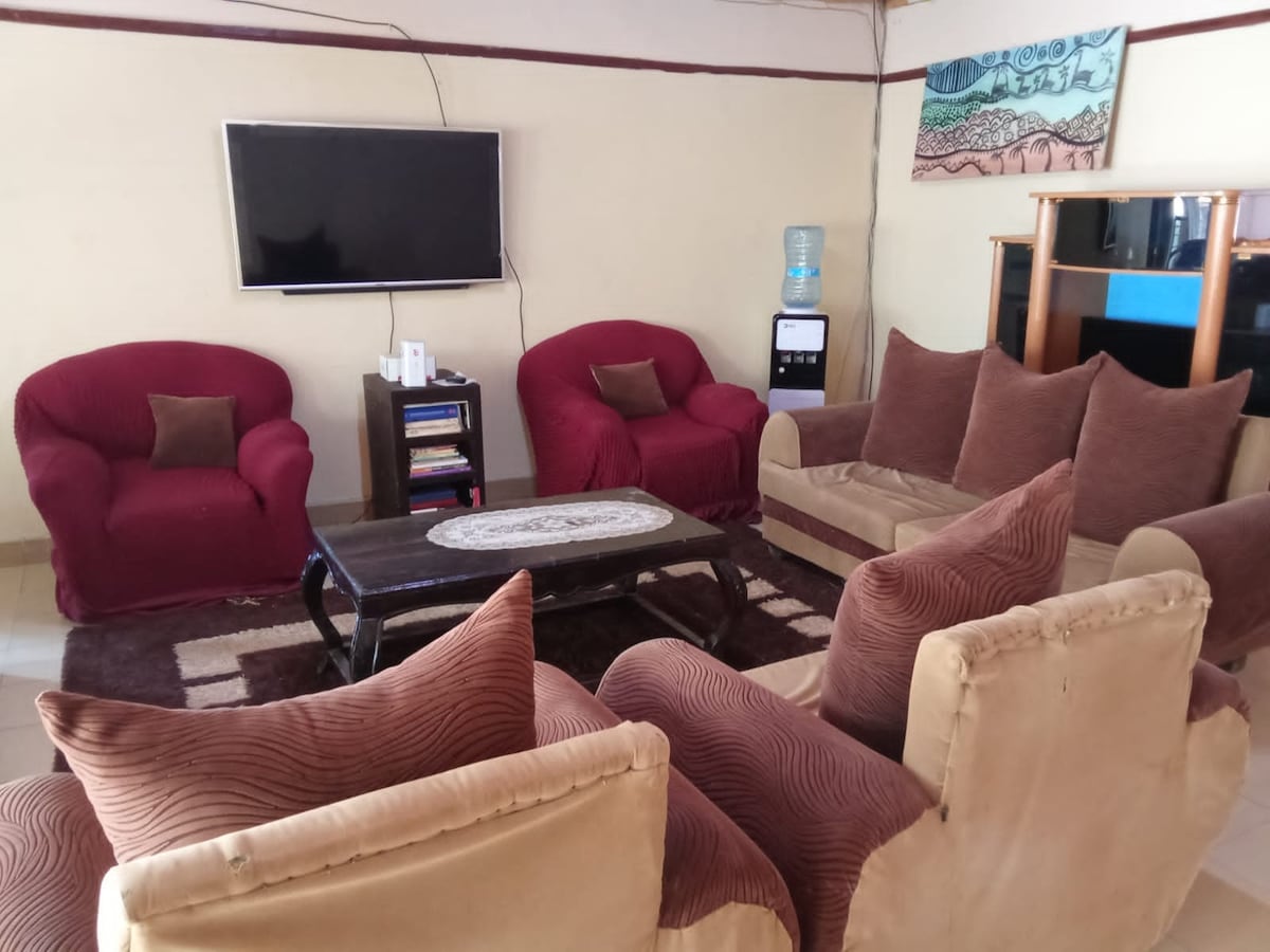 A spacious living area features a large coffee table surrounded by comfortable seating. Two red armchairs and a brown sofa set create a cozy arrangement. A television is mounted on the wall, and a water dispenser is visible in the background.