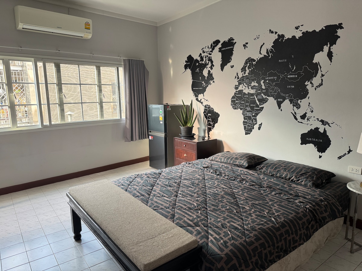 A spacious room features a king-size bed with a dark patterned coverlet and two pillows. A world map is displayed on the wall, enhancing the decor. A large window allows natural light, complemented by an air conditioning unit, and a refrigerator is positioned nearby.