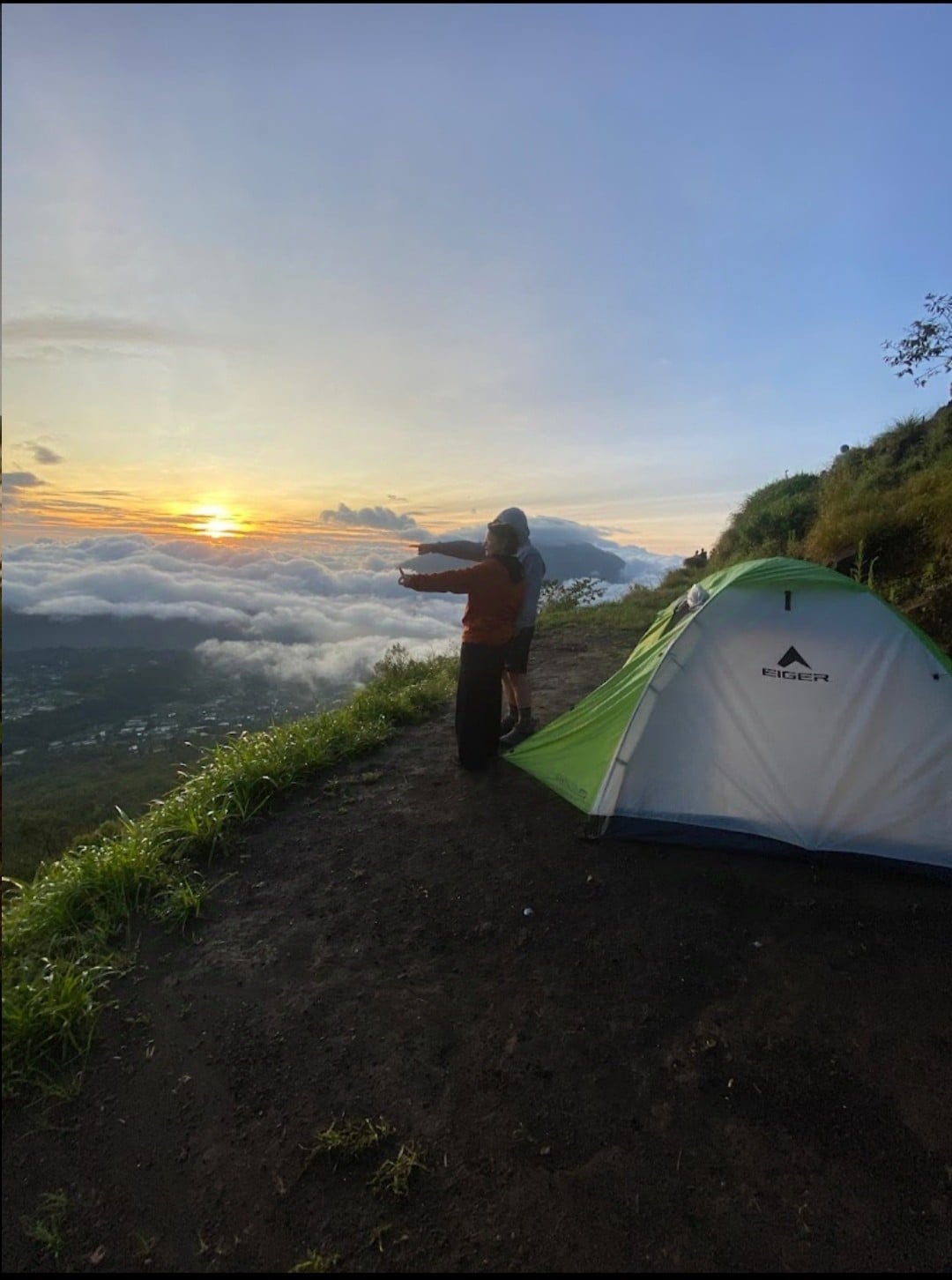 A tent sits on a grassy slope overlooking a sea of clouds during sunset. Two individuals stand nearby, facing the horizon where warm colors blend in the sky. The landscape features gentle hills, and the sky transitions from blue to golden tones, creating a serene backdrop.