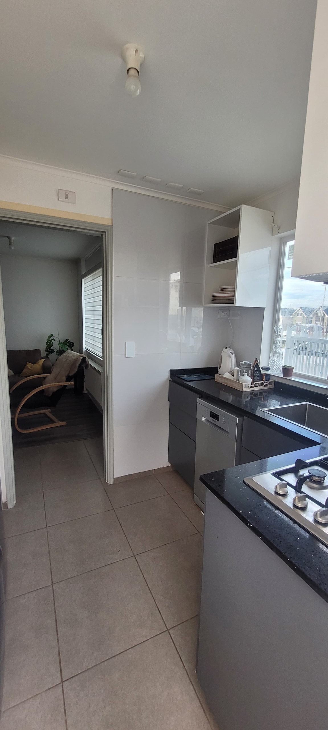 The kitchen area features sleek gray cabinetry and a black countertop. Various kitchen appliances, including a dishwasher and a stove, are visible. Sunlight enters through windows, illuminating the space. A doorway leads to a cozy seating area with a rocking chair and plants.