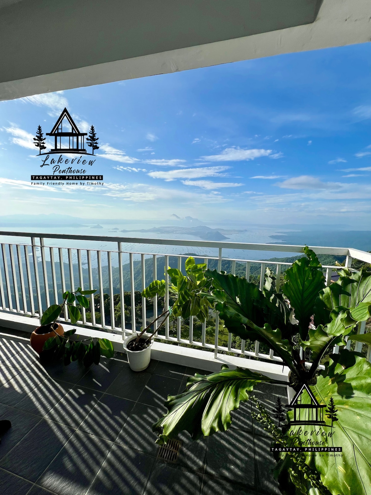 A spacious balcony offers sweeping views of Taal Lake and surrounding landscape. Decorative potted plants are positioned along the railing, with sunlight casting gentle shadows on the tiled floor. The serene atmosphere invites relaxation and enjoyment of the natural beauty.
