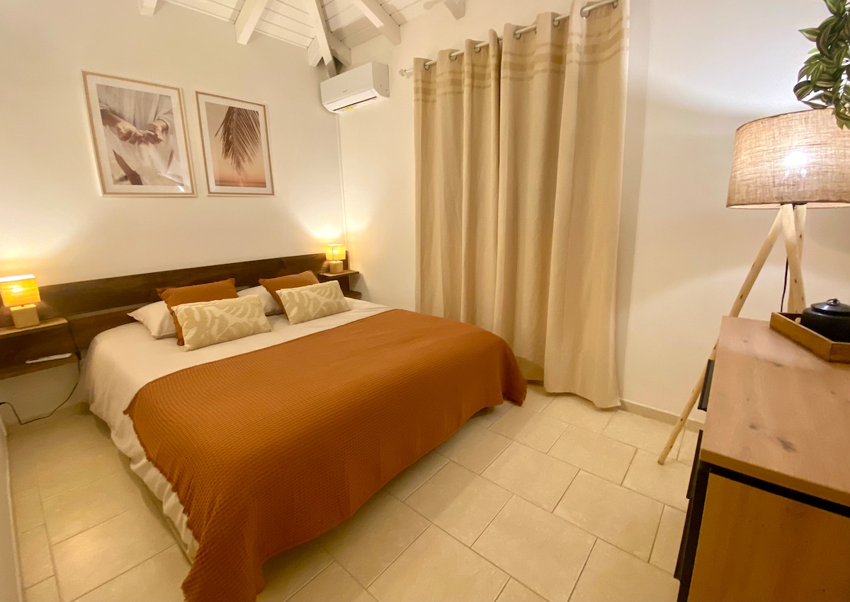 A cozy bedroom features a spacious bed dressed in warm, earthy tones. Soft lighting from bedside lamps creates a serene environment. Air conditioning is present, while light-colored curtains frame the window, allowing for privacy and gentle light diffusion. Neutral tile flooring contributes to the calming atmosphere.