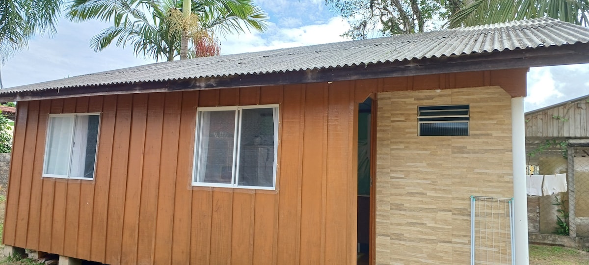 A wooden exterior of a single-story house is displayed, featuring large windows on both sides. The roof is corrugated metal, and a small vent is visible on the front wall. Lush greenery surrounds the structure, contributing to a natural environment.