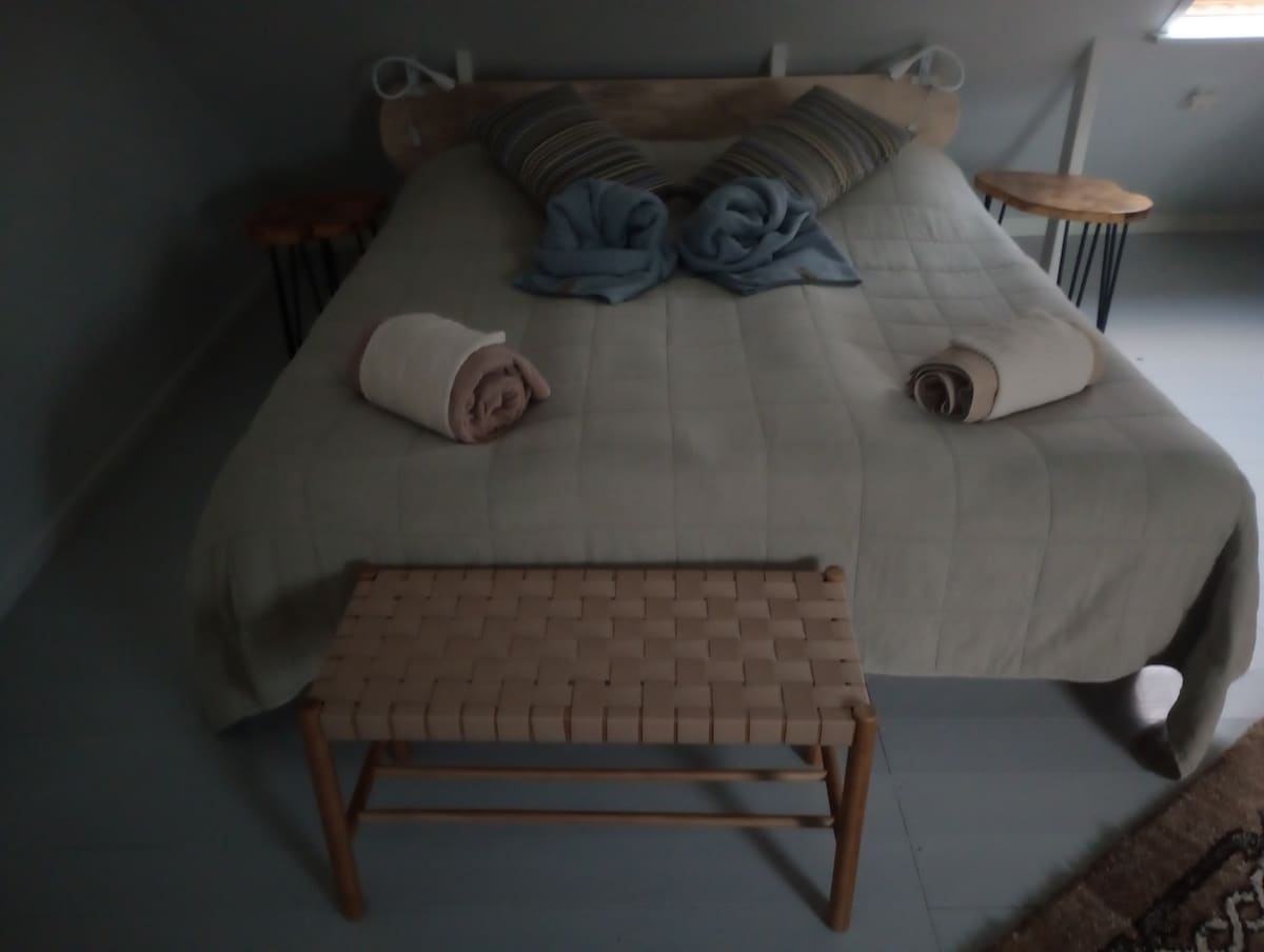 A spacious bed is centered in the room, adorned with a neutral-colored quilt. Two striped pillows and rolled towels add a touch of comfort. Wooden side tables sit on either side, and a woven bench is placed at the foot of the bed, completing the arrangement.
