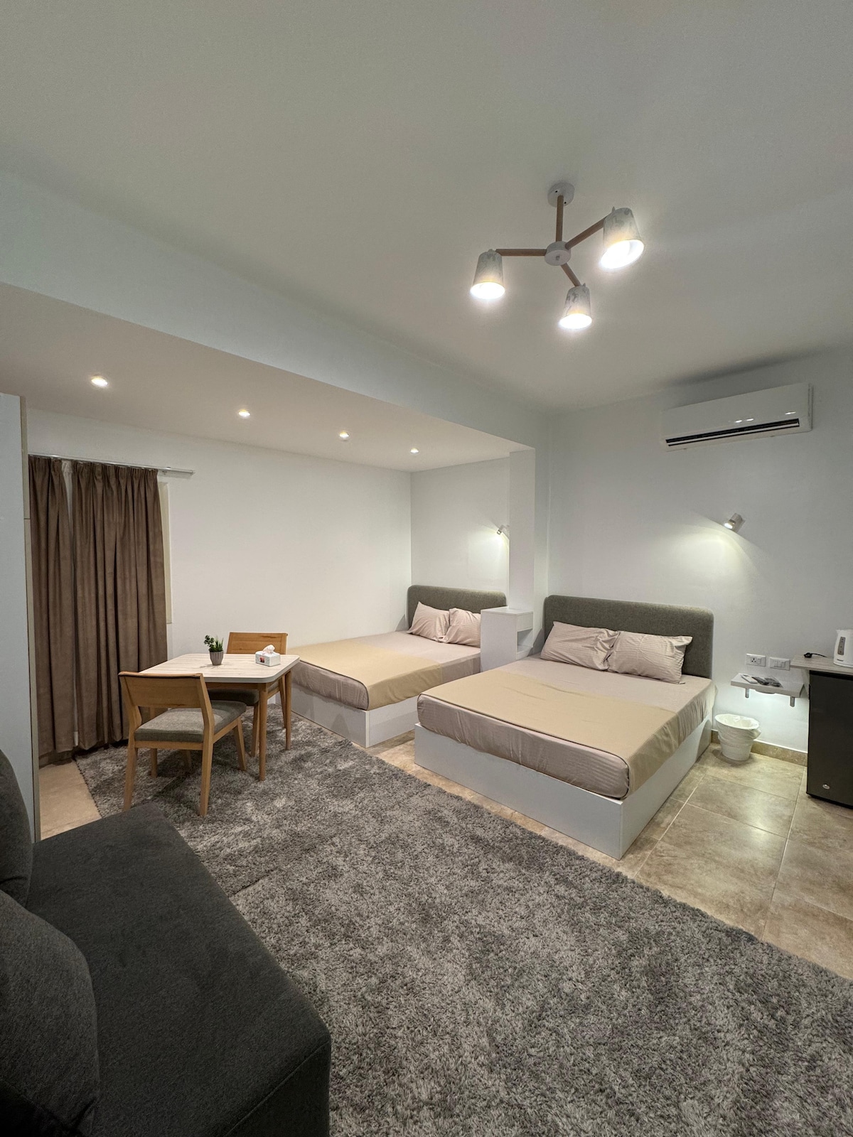 The room features two beds with soft linens, complemented by a cozy seating area with a small table and chairs. Soft lighting from the ceiling and a nearby wall creates a warm ambiance. A dark curtain frames a window, enhancing privacy.