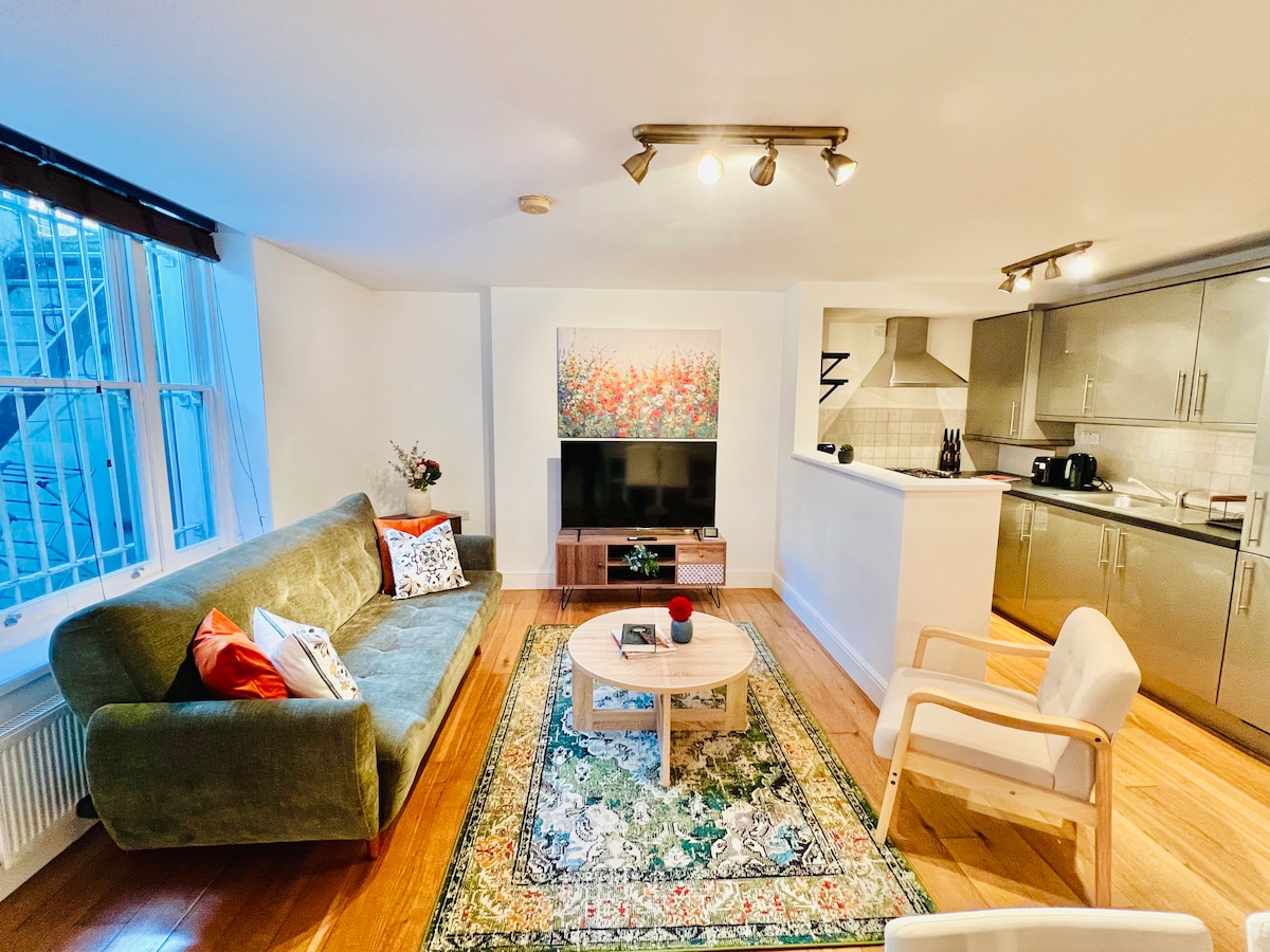 A comfortable living space is presented, featuring a green sofa adorned with accent pillows. A large piece of artwork with floral themes is displayed above the TV unit. A wooden coffee table sits on an area rug, while the kitchenette is visible in the background.