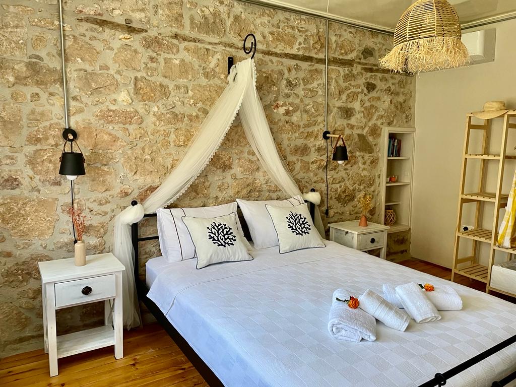 A spacious sleeping area features a stone wall that adds texture to the room. A large bed is adorned with soft linens, complemented by two bedside tables. Decorative pillows and hand towels are neatly arranged, while a woven pendant light hangs above, enhancing the serene ambiance.