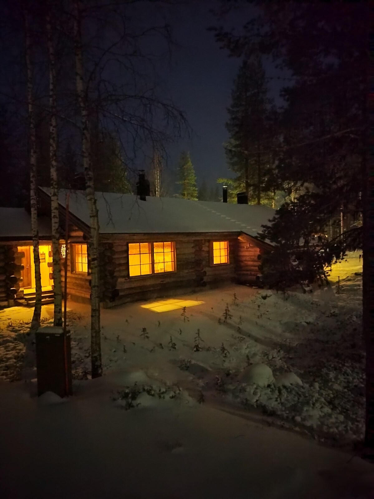 A log cabin is nestled among snow-covered trees, with warm light emanating from illuminated windows. The snowy ground reflects the soft glow, creating a serene evening atmosphere. The cabin's rustic architecture is highlighted against the natural backdrop of a wintry landscape.