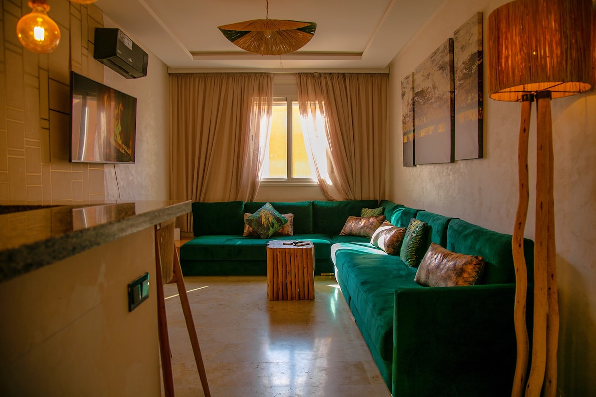 A spacious living area features a modern design with a vibrant green sectional sofa and a natural wood coffee table. Soft curtains frame the window, allowing natural light to brighten the room. A unique pendant light and a floor lamp add warmth to the space.