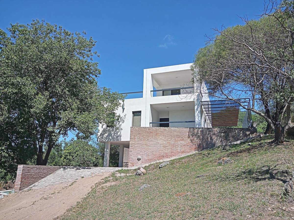 The modern two-story structure is situated on a slight hillside, surrounded by greenery. Large windows invite natural light, and a balcony extends from the upper floor, offering views of the landscape. Natural stone and earth tones complement the exterior design.