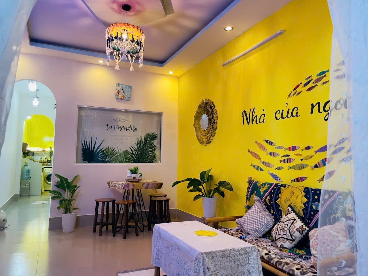 A vibrant living space features a yellow accent wall adorned with decorative fish designs. A cozy seating area includes a low table framed by two chairs. Decorative plants are placed strategically, while soft lighting is provided by a stylish overhead fixture.