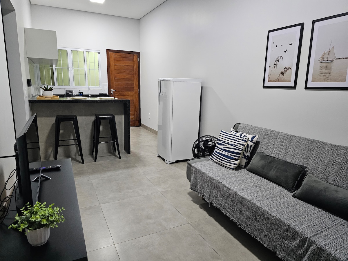 The living area features a cozy gray sofa with decorative cushions, complemented by a flat-screen TV on a black console. The open kitchen includes a refrigerator and a bar with two stools, creating a functional space for relaxation and dining.