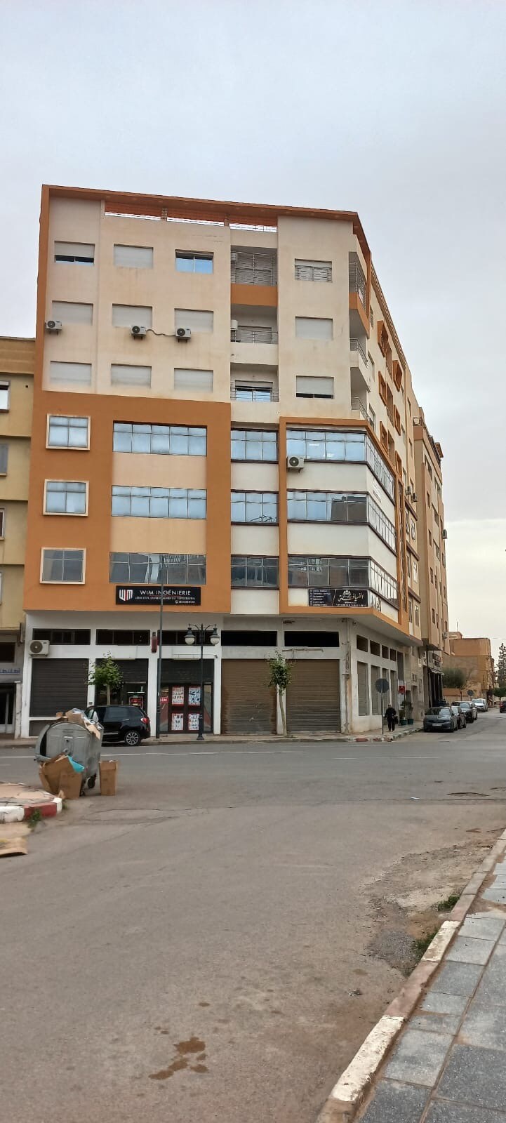 A modern multi-story building is observed from the street, showcasing a facade with large windows and a combination of beige and orange tones. Various air conditioning units are mounted on the exterior. Nearby, shops and parked cars can be seen along the road.