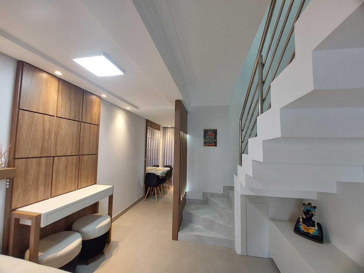 A well-lit interior space features a modern staircase with a sleek design. To the left, a wooden accent wall complements a stylish dining area with round chairs. A minimalist table is positioned near the entrance, creating an open and inviting layout.