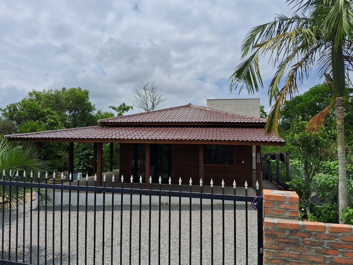 The exterior of a wooden house is showcased under a cloudy sky. A sloped, tiled roof covers a spacious porch. Surrounding greenery adds a touch of nature, while a low, brick wall with metal fencing defines the boundary of the property.