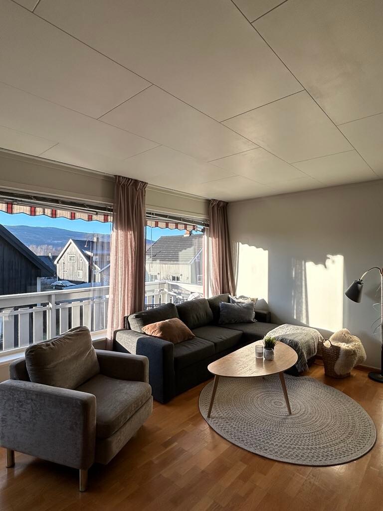 Natural light brightens a cozy living area, featuring a comfortable sofa and two armchairs. A round coffee table sits on a textured rug. Large windows with light curtains frame a view of the surrounding buildings and landscape, inviting a sense of openness.