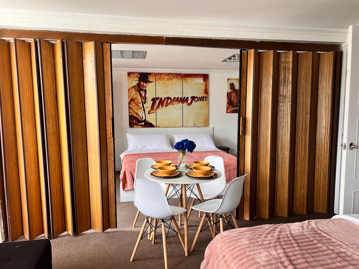 The suite features a partition with wooden sliding doors, separating the sleeping area from a small dining space. The room displays a colorful 'Indiana Jones' movie poster. A round table is set for four, complemented by white chairs and a vibrant blue flower arrangement.