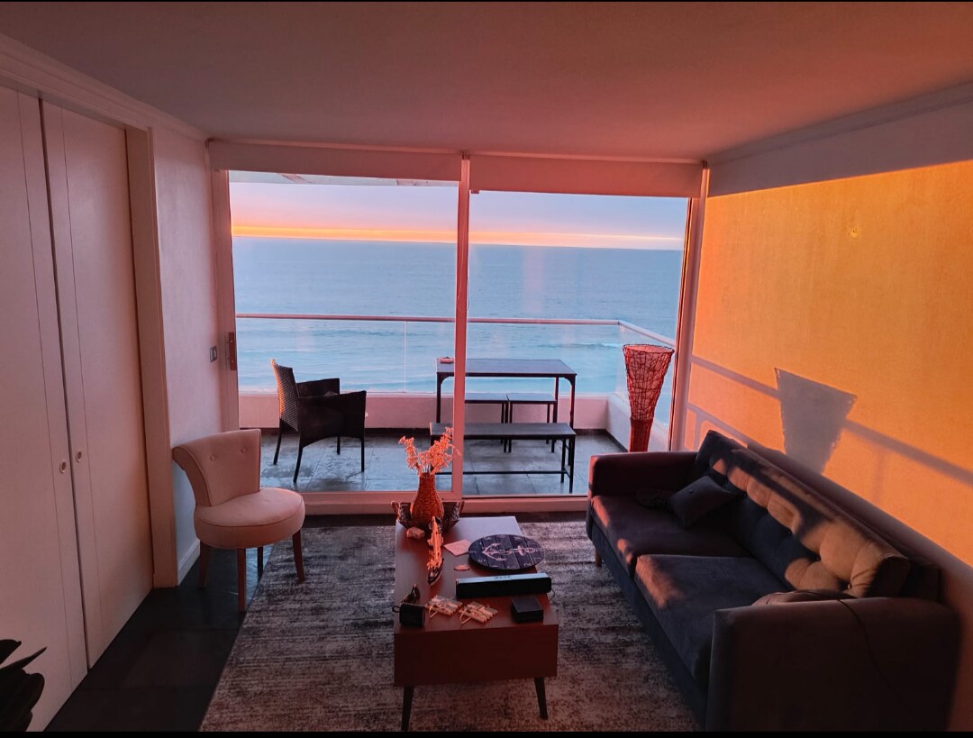 A bright living area features large glass windows that offer sweeping ocean views at sunset. A plush sofa and side chair are arranged around a wooden coffee table, with soft lighting creating a serene ambiance. The space is complemented by a neutral-toned rug.