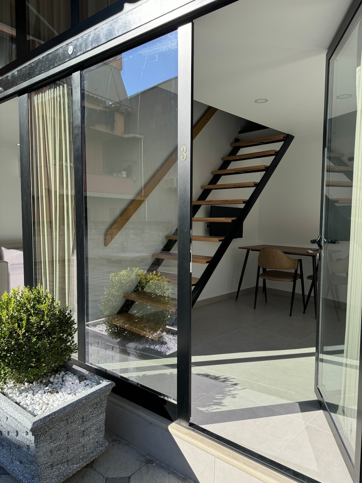A modern entryway features sleek glass doors leading to a minimalist staircase with wooden steps. A small dining table and chair are visible in the background, while neatly arranged planters with greenery add a touch of nature to the welcoming space.