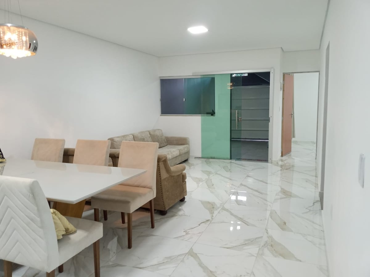 A bright dining area features a long table with light-colored chairs and a cozy sofa nearby. The glossy marble floor reflects the light from the ceiling fixture, while glass doors lead to a private outdoor space.