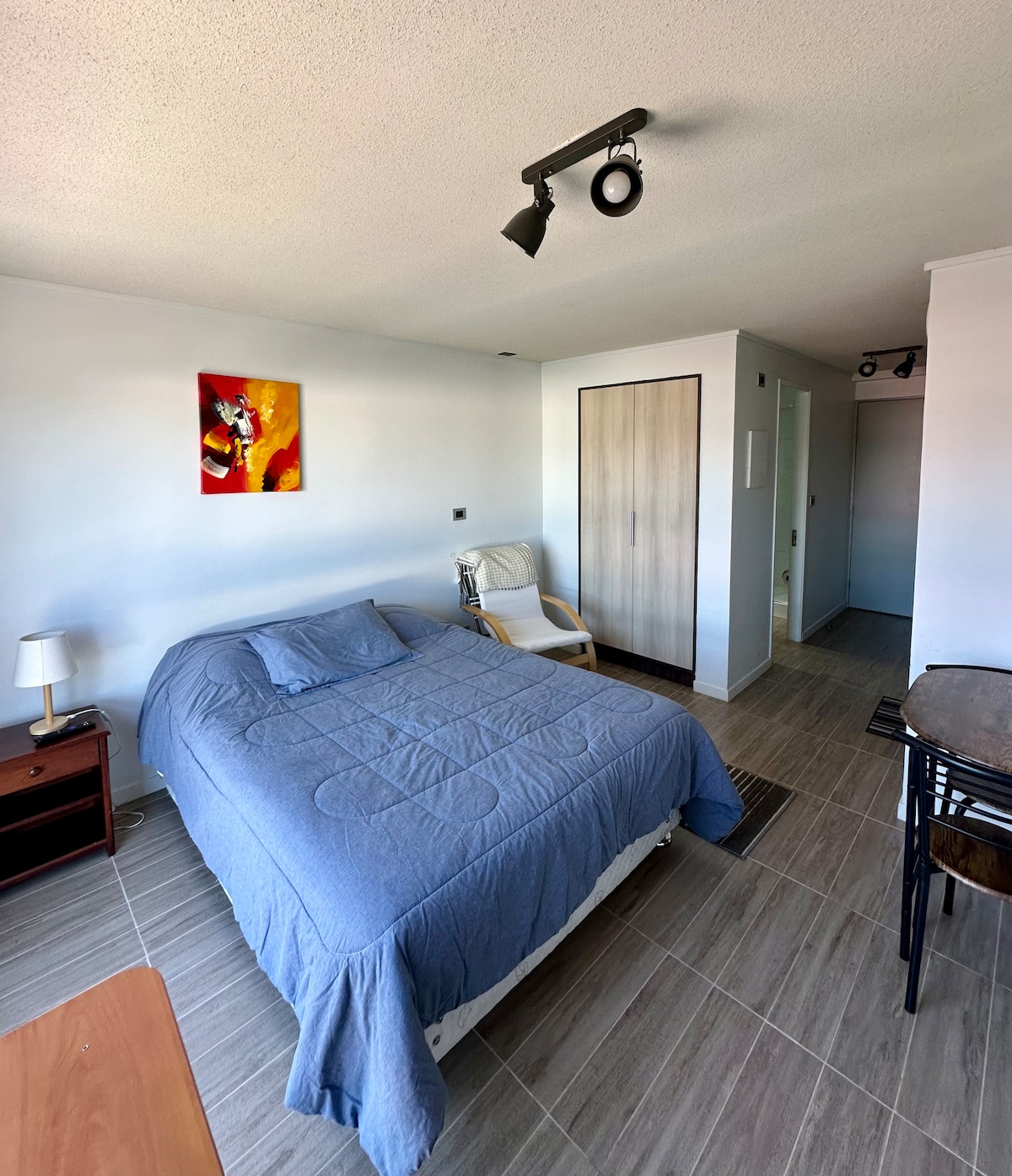 A studio living space showcases a comfortable bed with a blue quilt, accompanied by a bedside table and a lamp. A piece of artwork adds a splash of color to the wall. A closet is present, and light filters in from a nearby window.