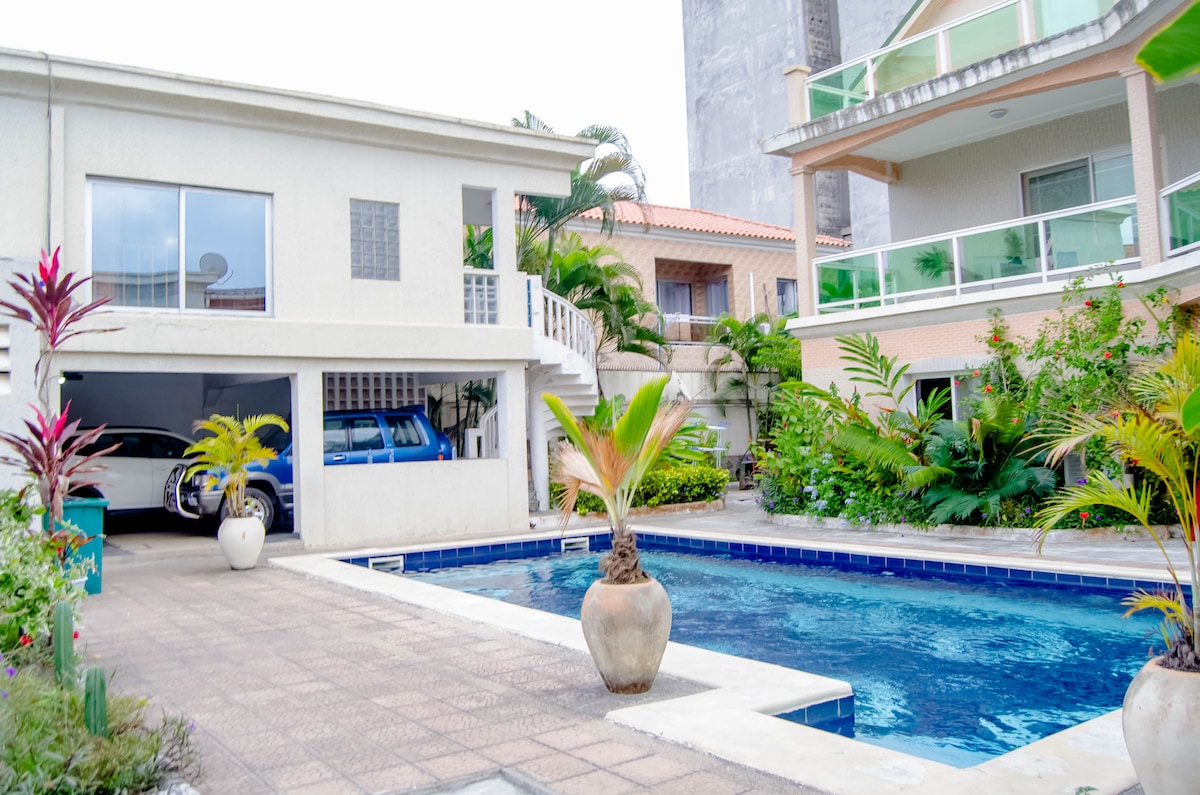 A modern villa is surrounded by lush greenery and features a private pool. Spacious outdoor areas are accentuated by potted plants. Two vehicles are parked in the garage, with a staircase leading to the upper level visible in the background.