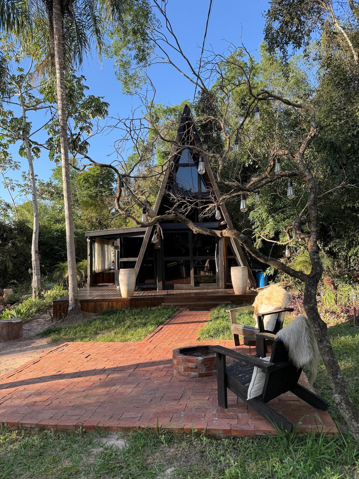 An A-frame chalet is set amidst lush greenery, showcasing large glass windows. A brick path leads to a small seating area with chairs and decorative planters. Surrounding trees add to the natural ambiance, creating a serene outdoor space.
