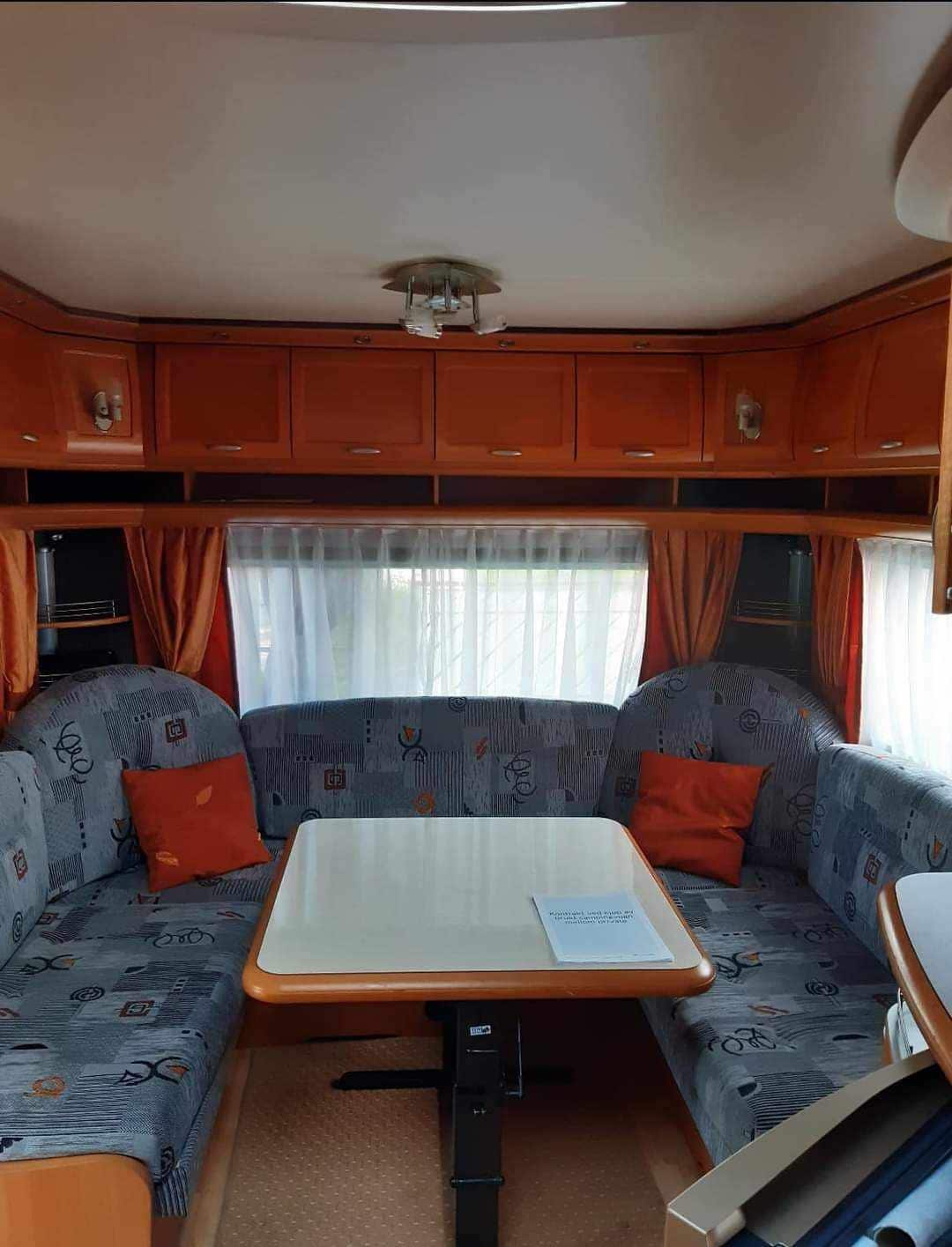A functional seating area is displayed, featuring a light-colored table surrounded by cozy, curved benches. The cushions are adorned with subtle patterns, and bright orange pillows add a pop of color. Natural light filters through a curtain-covered window, illuminating the interior.
