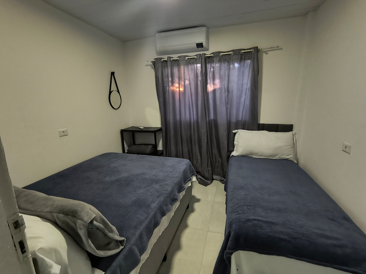 A simple sleeping area features two beds with blue blankets, providing a calm ambiance. A dark curtain frames the window, allowing soft light to filter in. A compact desk stands against one wall, adding functionality to the space.