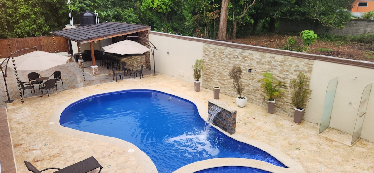 A refreshing pool with a waterfall feature occupies the center of the outdoor space. Two shaded seating areas with umbrellas are visible, accompanied by a stone wall that enhances the serene environment. Lush greenery surrounds the area, creating a calm and inviting retreat.
