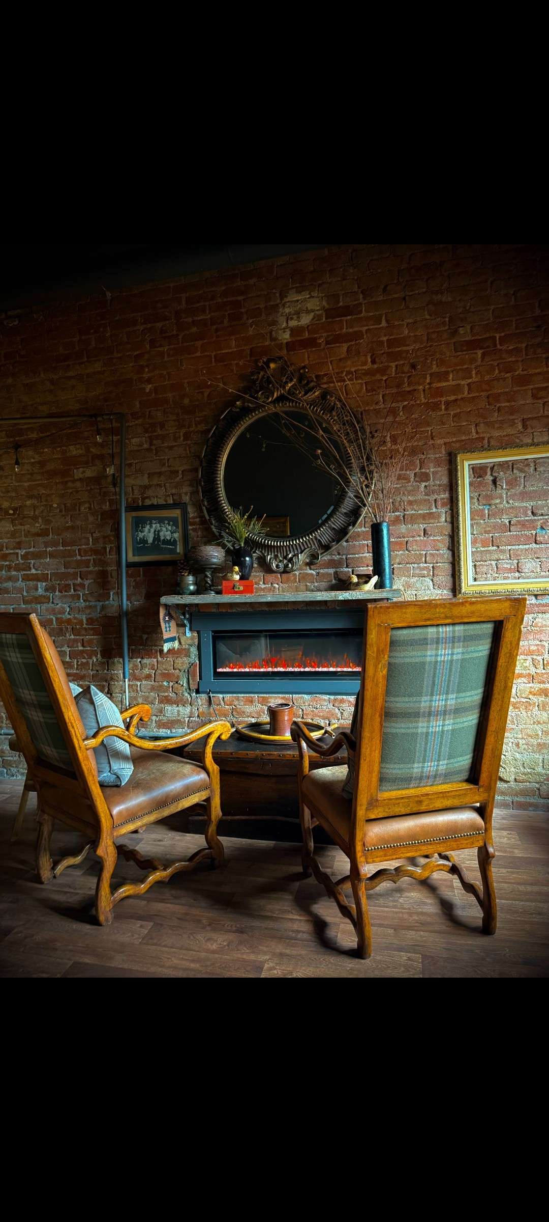 A cozy seating area features two antique wooden chairs positioned around a small table. A contemporary electric fireplace is set against a rustic brick wall, complemented by a large round mirror above. Decorative items are arranged on the mantel, enhancing the space's character.