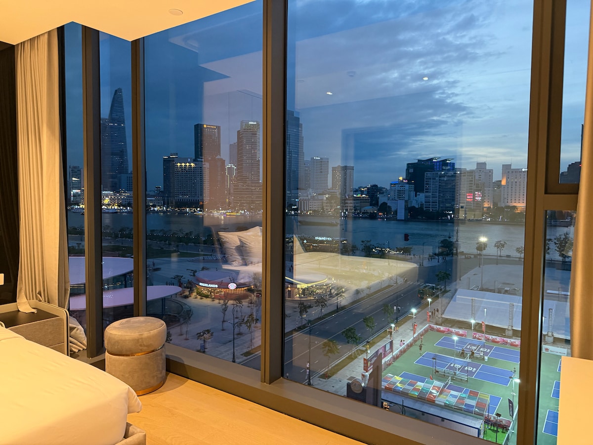 A spacious room features floor-to-ceiling windows showcasing a stunning view of the city skyline and river. The gentle evening light reflects off the buildings, creating a serene ambiance. The scene includes a tennis court and beautifully landscaped areas below.