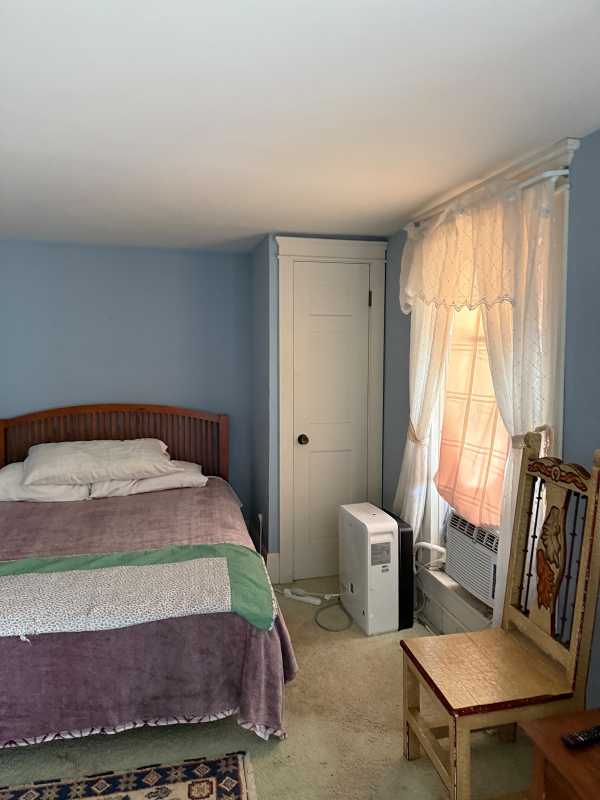 A cozy bedroom features a sizable bed with layered blankets in soft colors. A chair with a decorative backrest is positioned beside the bed. An air conditioning unit stands near a window draped with light, airy curtains, enhancing the serene environment.