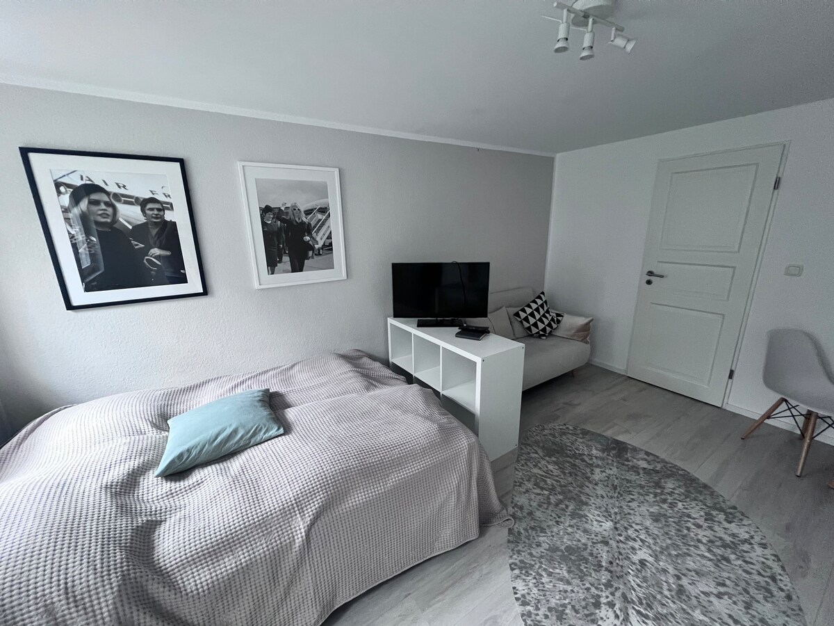 A well-lit room features a spacious bed covered with a neutral-toned blanket. Beside it, a small sofa is positioned next to a modern white TV stand. Art pieces frame the wall, and light-colored flooring enhances the sense of space.