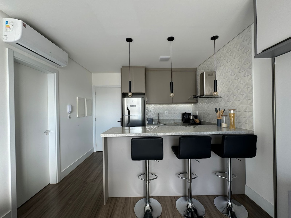 A modern kitchen area features a sleek countertop with three black bar stools. Pendant lights hang above, illuminating the space. Stainless steel appliances and minimalist cabinetry are visible, while a textured wall adds visual interest.