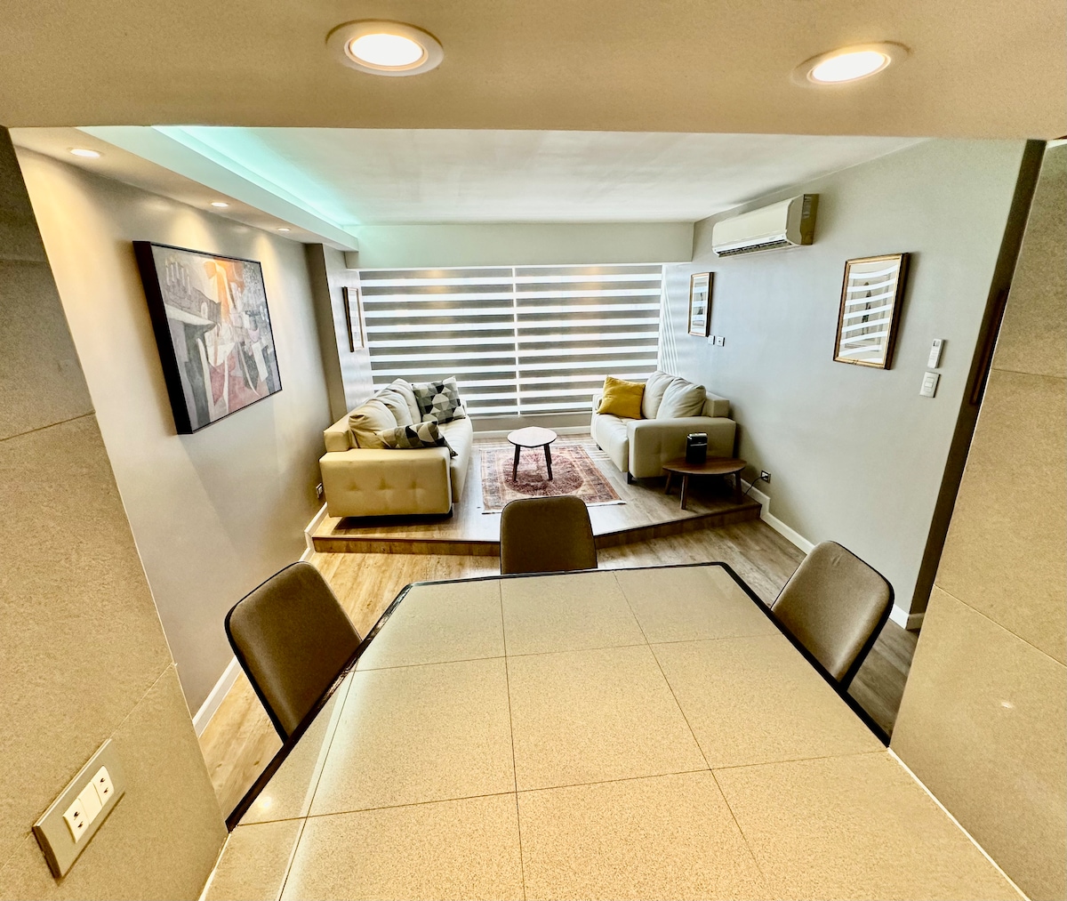 The living area features two comfortable seating arrangements, one adorned with a soft throw and decorative pillows. A small round coffee table sits in the center, and natural light filters through modern blinds. A sleek dining table is positioned in the foreground.