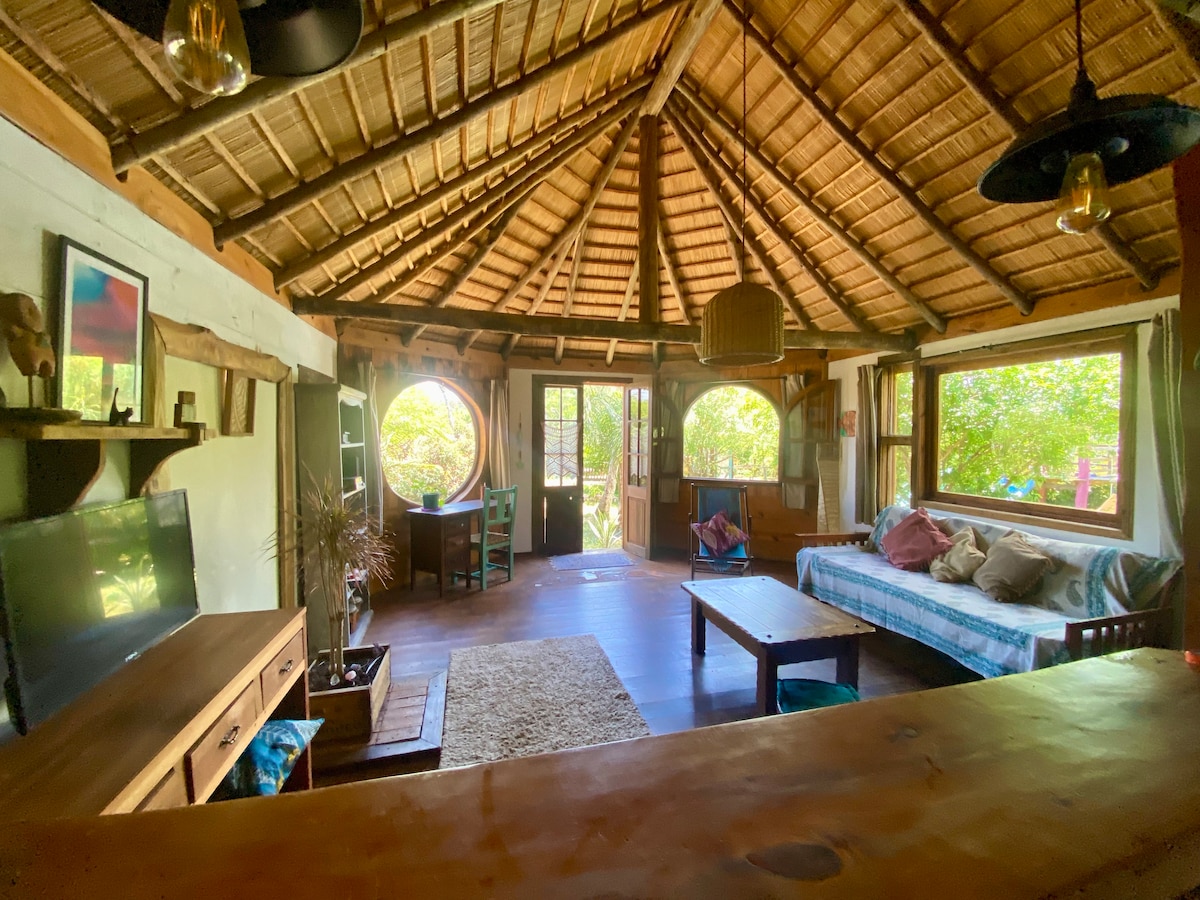 A circular living area features a high, thatched roof made of natural materials. Large windows provide ample natural light, and cozy seating is arranged around a woven rug. A wooden table and entertainment unit create a welcoming space for relaxation and connection.