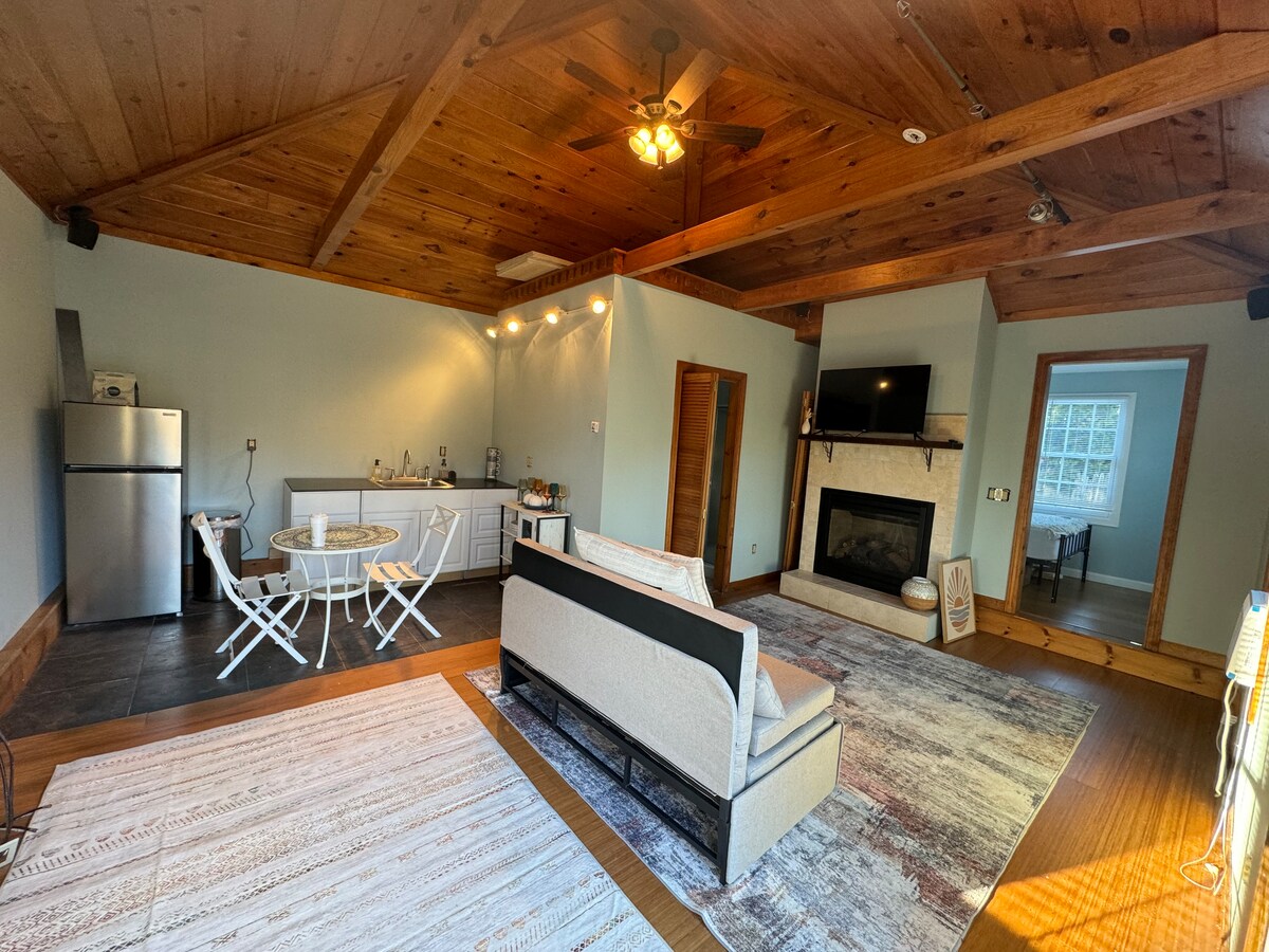 The open living space is highlighted by wooden ceiling beams and features a kitchenette with stainless steel appliances. A dining area equipped with a round table and two chairs is present. A cozy loveseat faces a gas fireplace, adding a focal point to the room.