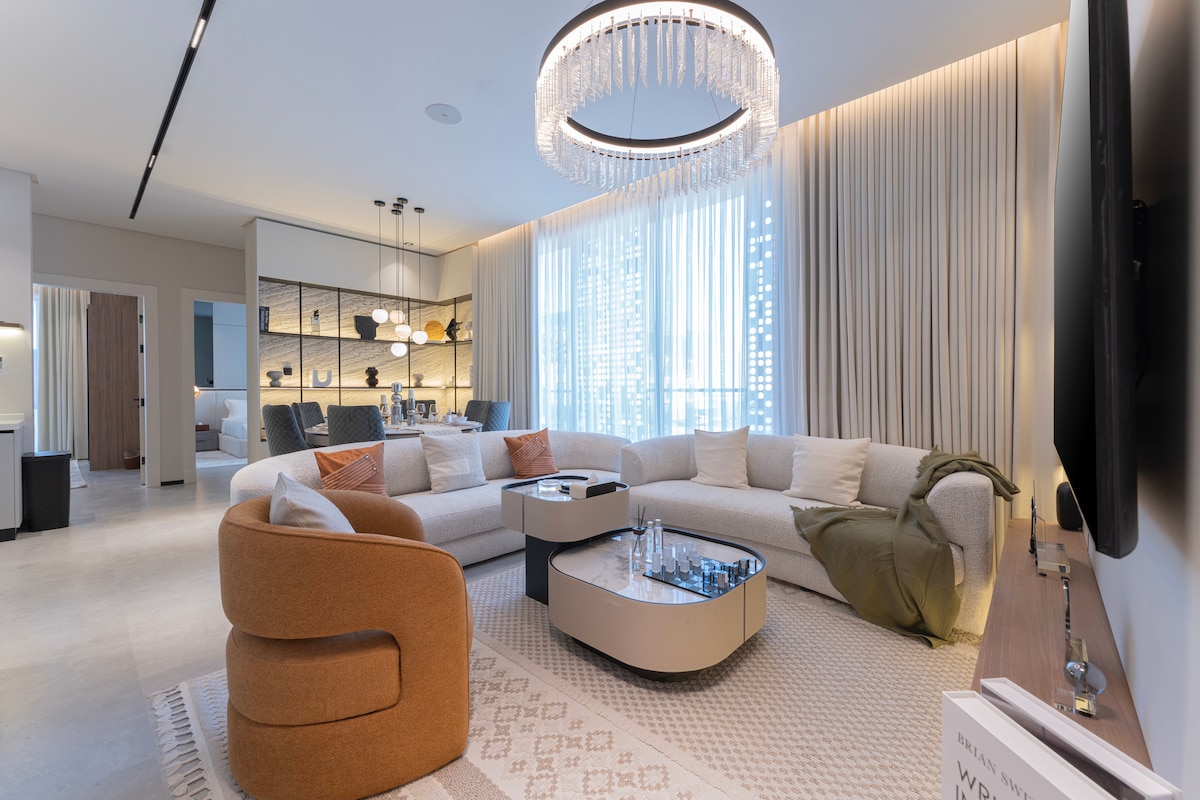 A spacious living area features a large, curved sectional sofa with accent pillows, positioned around a sleek coffee table. A modern chandelier hangs overhead, and floor-to-ceiling windows are adorned with soft drapes, allowing natural light to fill the space. A cozy throw is draped over the arm of the sofa.