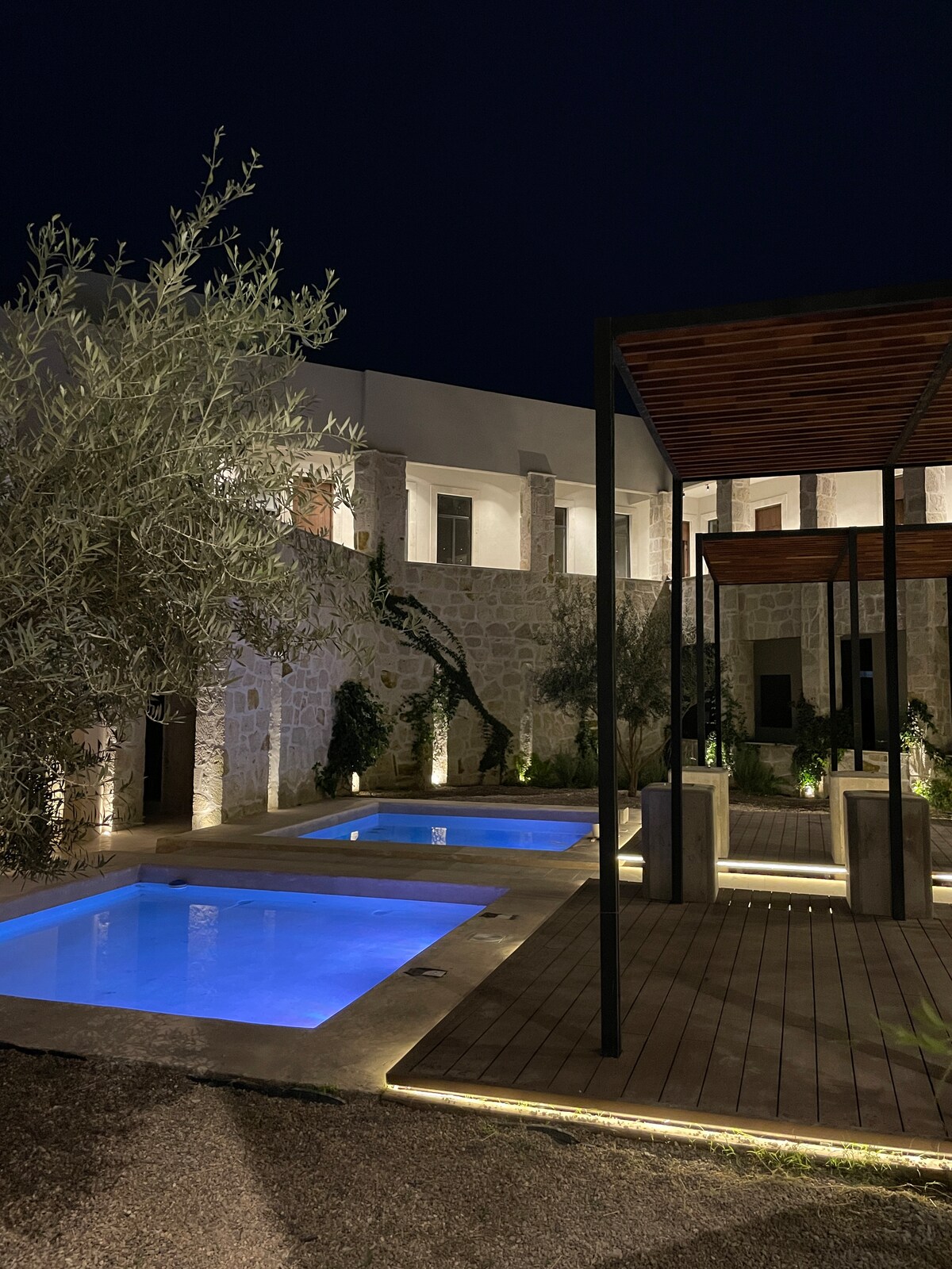 Two illuminated plunge pools are surrounded by a stone patio, with greenery and low lighting enhancing the evening scene. A wooden deck provides a space for relaxation, framed by the structured layout of the building, creating a serene and inviting atmosphere.