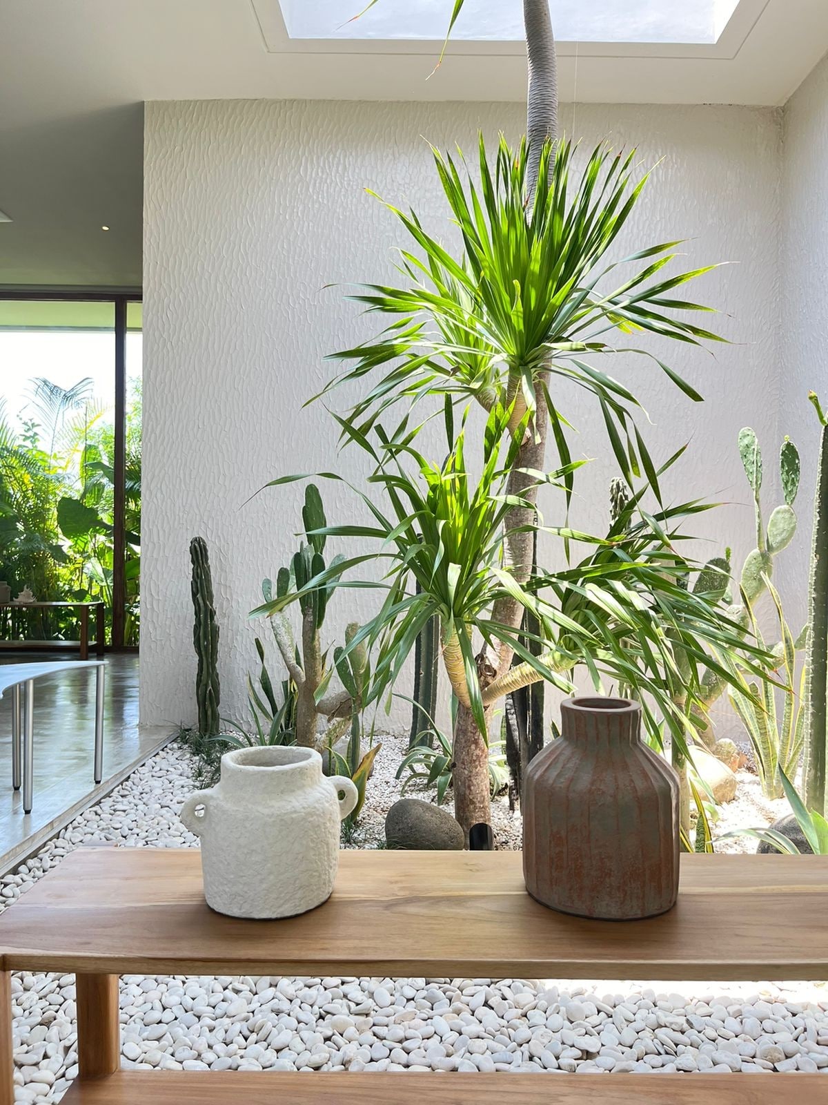 A wooden bench displays two decorative vases, one textured and white, the other terracotta. Behind them, lush green plants and cacti can be seen in a bright, peaceful indoor garden space, creating a serene atmosphere.