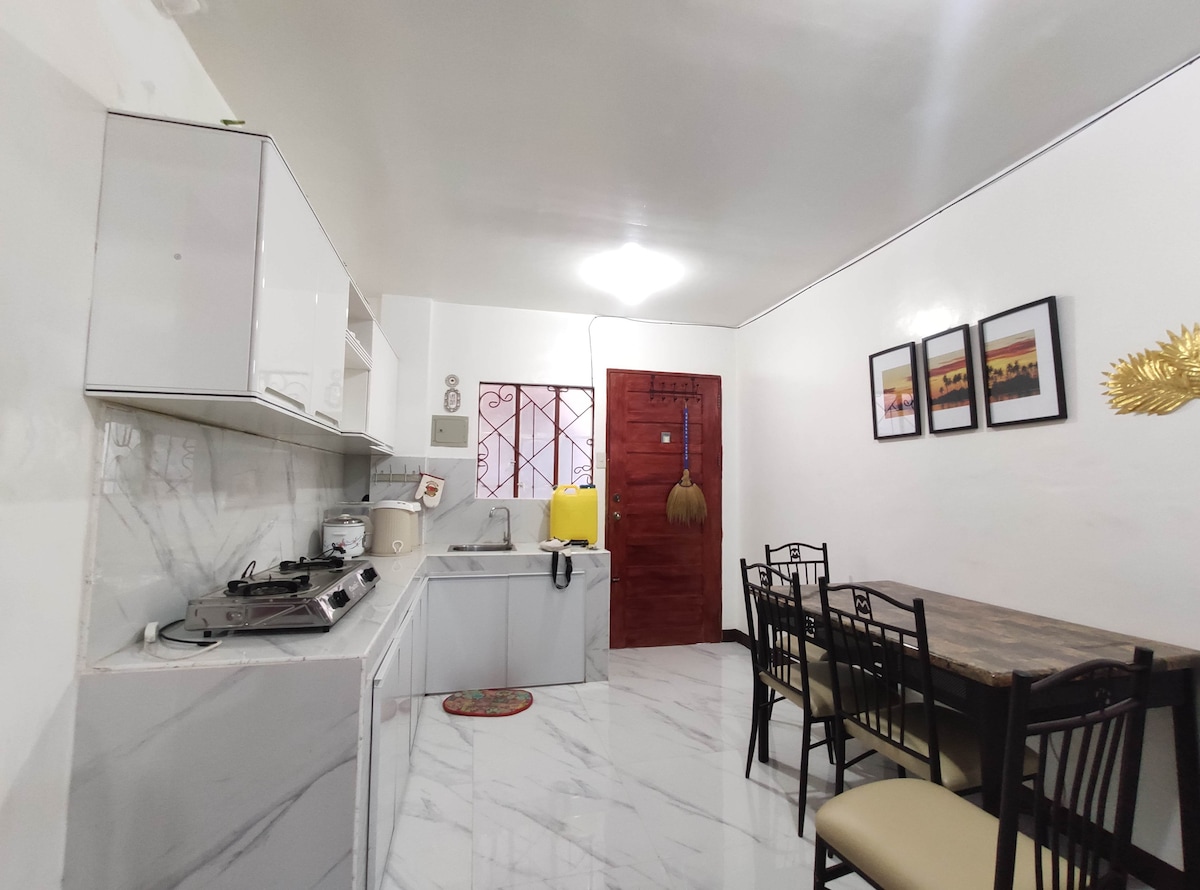 A functional kitchen area is presented with modern cabinets and sleek marbled countertops. Cooking appliances are neatly arranged, while a dining table with chairs invites comfortable meals. Bright artwork adorns the wall, enhancing the space's character.