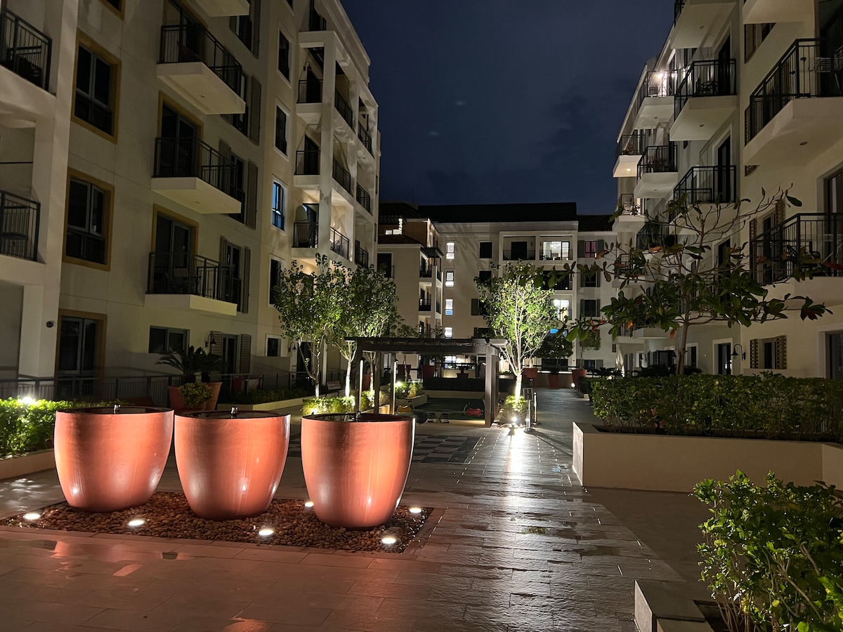 An evening view of the courtyard features three large, lit planters positioned in a neatly landscaped area. Surrounding buildings reflect gentle lighting, enhancing the tranquil atmosphere. Pathways are visible, leading through the greenery towards a central gazebo, with additional landscaping offering soft illumination.