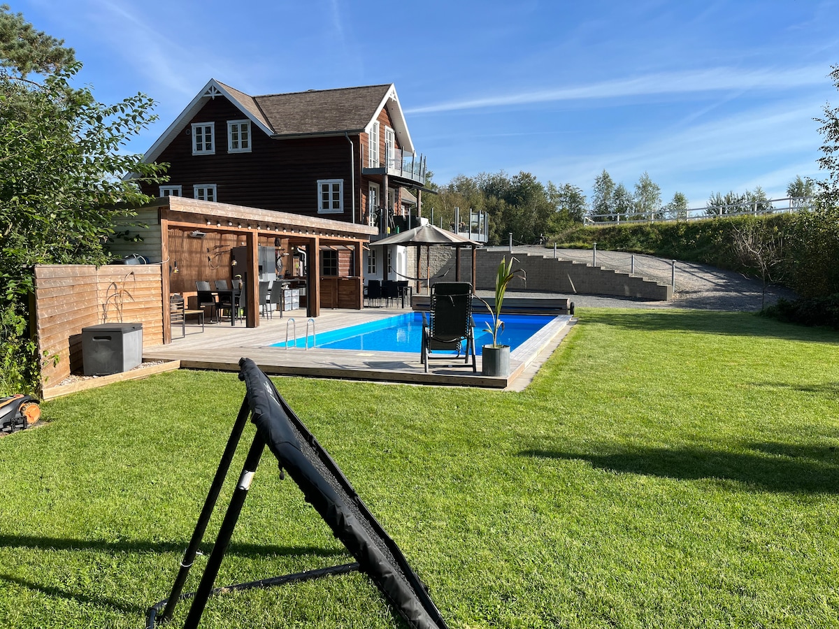 A serene outdoor space features a swimming pool surrounded by a wooden deck. The grassy area is bordered by trees, providing privacy. A spacious building with multiple stories is visible in the background, showcasing a blend of natural and residential elements.