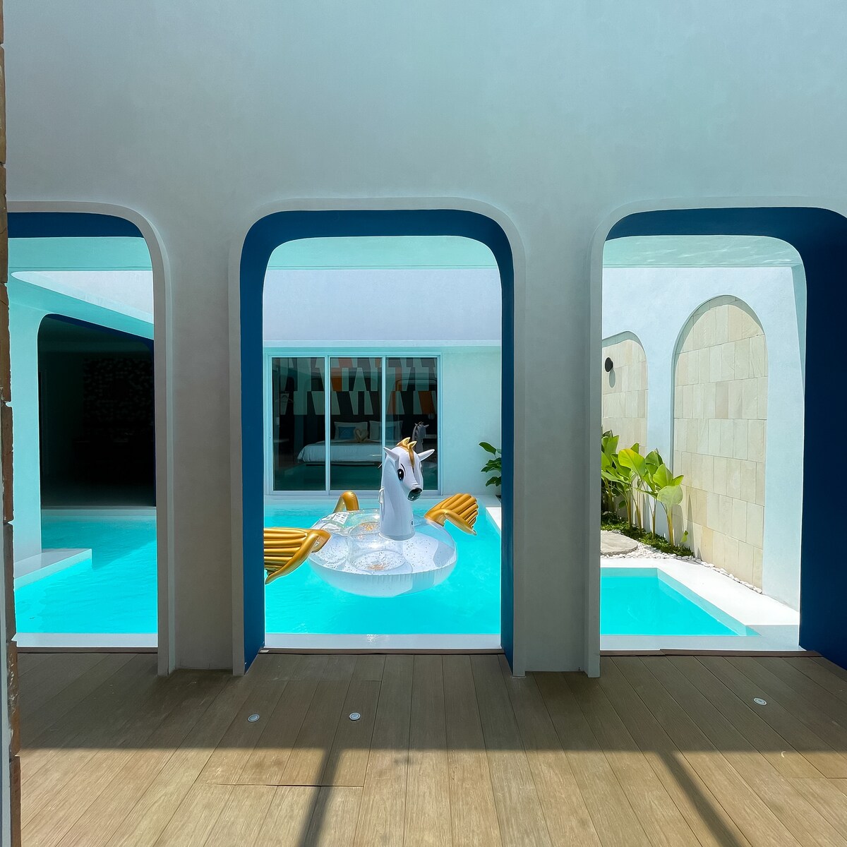 A view of the private pool is framed by arched openings in the villa's walls. A vibrant unicorn float is positioned in the turquoise water, alongside two yellow lounge chairs arranged for relaxation. Sunlight filters through the structure, emphasizing the pool's inviting clarity.