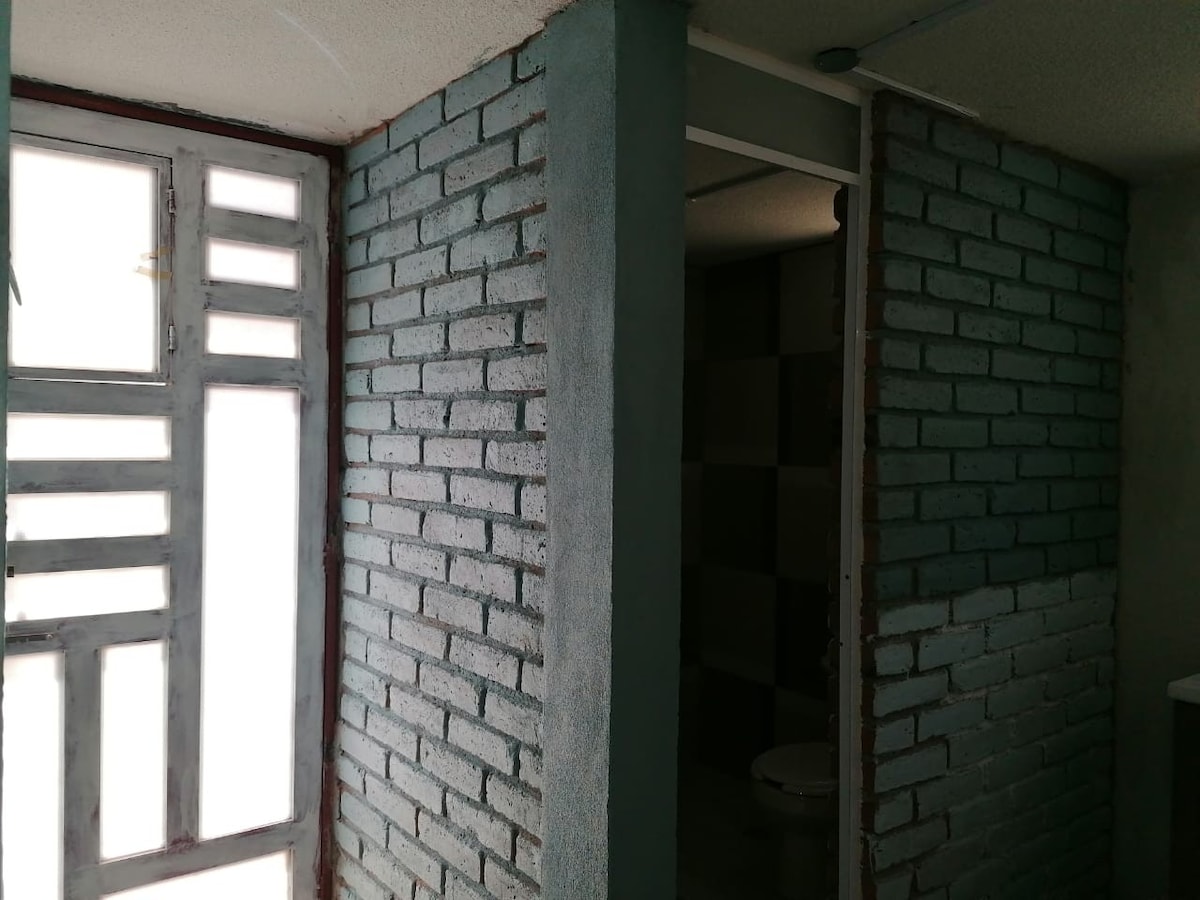 The interior features brick walls painted in a muted blue tone, complemented by a large window allowing natural light to filter in. A door leading to a bathroom is visible on the right, providing access to private facilities.