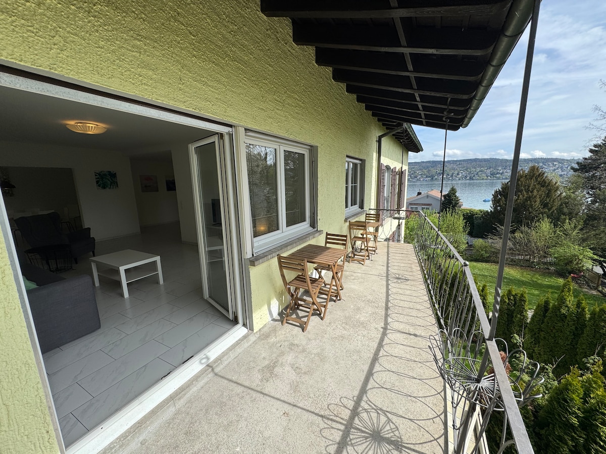 A spacious balcony is visible, featuring a seating area with two folding chairs and a small table. Sliding glass doors lead into the living area, allowing for natural light. Lake Zurich can be seen in the background, complemented by distant mountains.