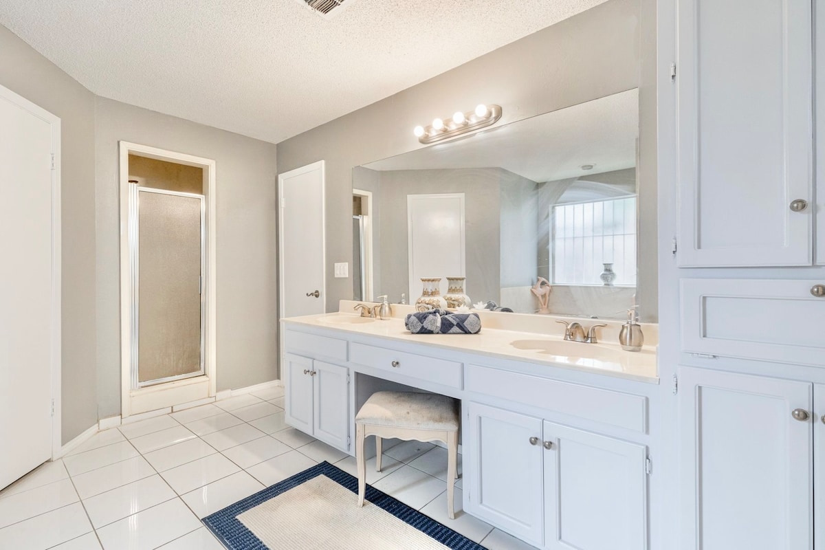 A bright bathroom is showcased, featuring a spacious double vanity with ample storage beneath. A large mirror reflects natural light, enhancing the clean aesthetic. A shower area and neutral tile flooring provide a functional and inviting space, complemented by simple decorative accents.