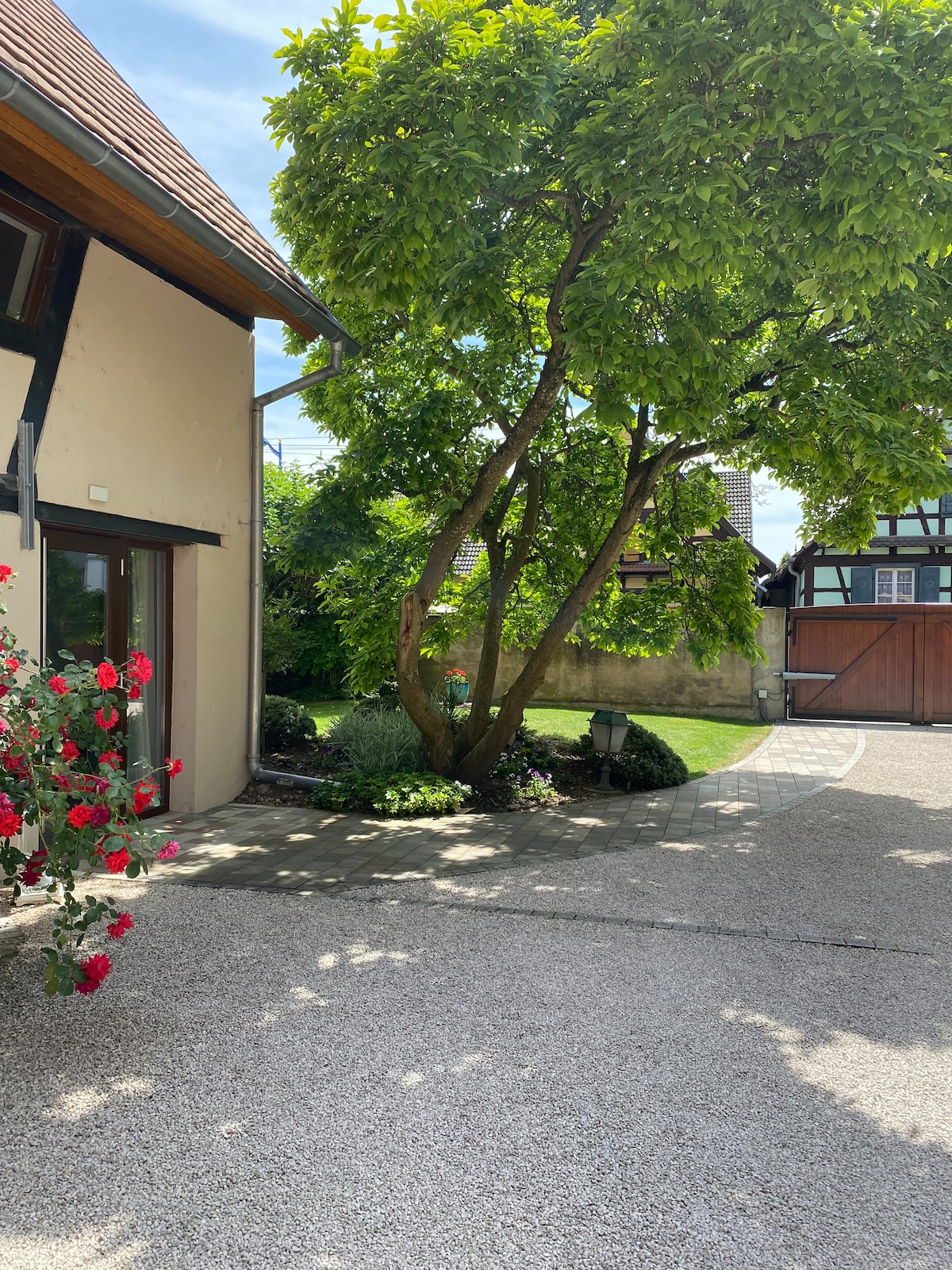 A well-maintained courtyard is featured, surrounded by lush greenery and blooming rose bushes. A mature tree provides shade, while the gravel path leads towards an inviting entrance. The walls of the structure show a soft hue, complementing the natural elements present.